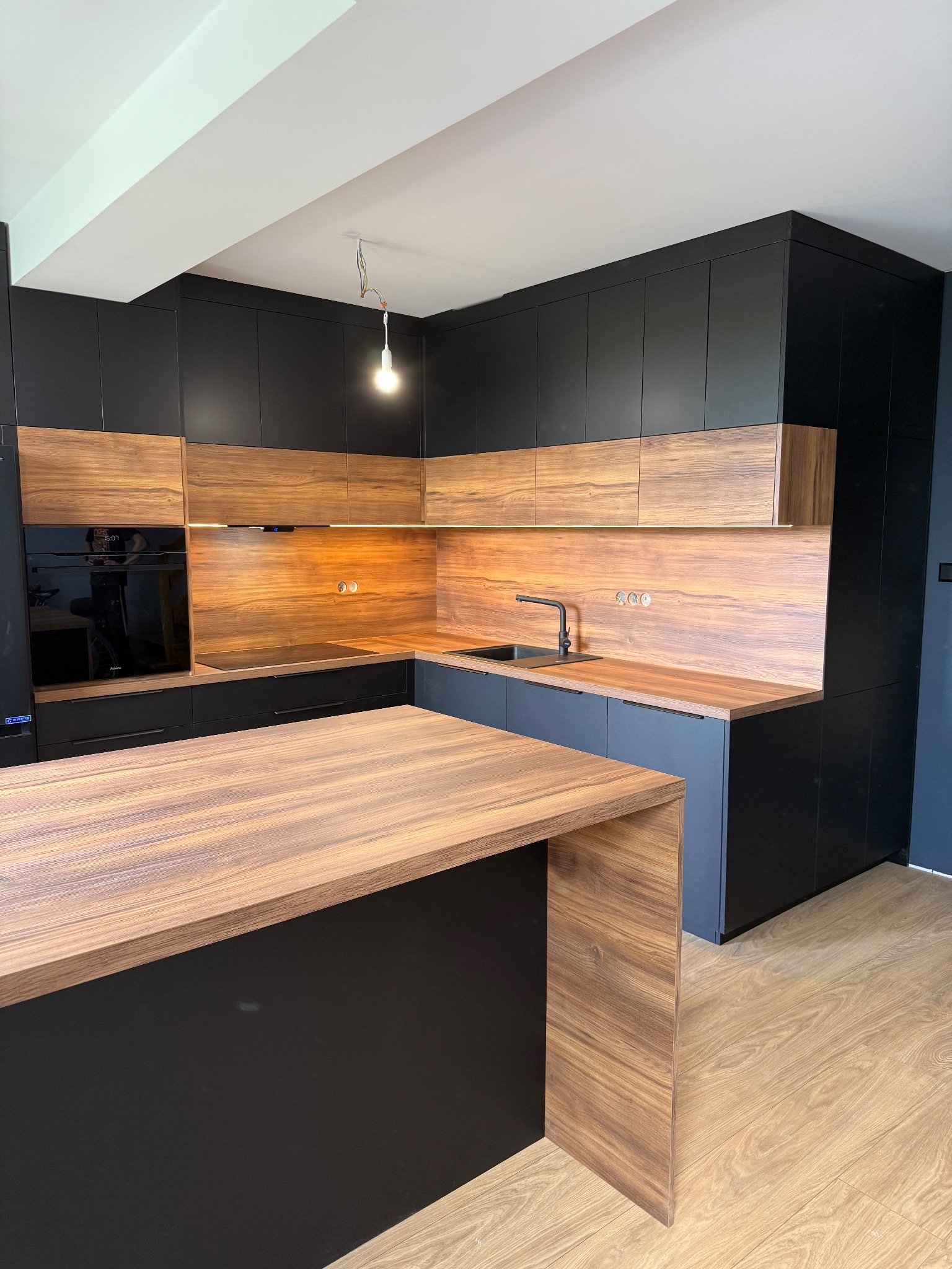 Nowoczesna kuchnia z czarnymi szafkami i drewnianymi blatami. Wyspa kuchenna z blatem w kolorze drewna. Widoczne oświetlenie podszafkowe i sprzęt AGD.