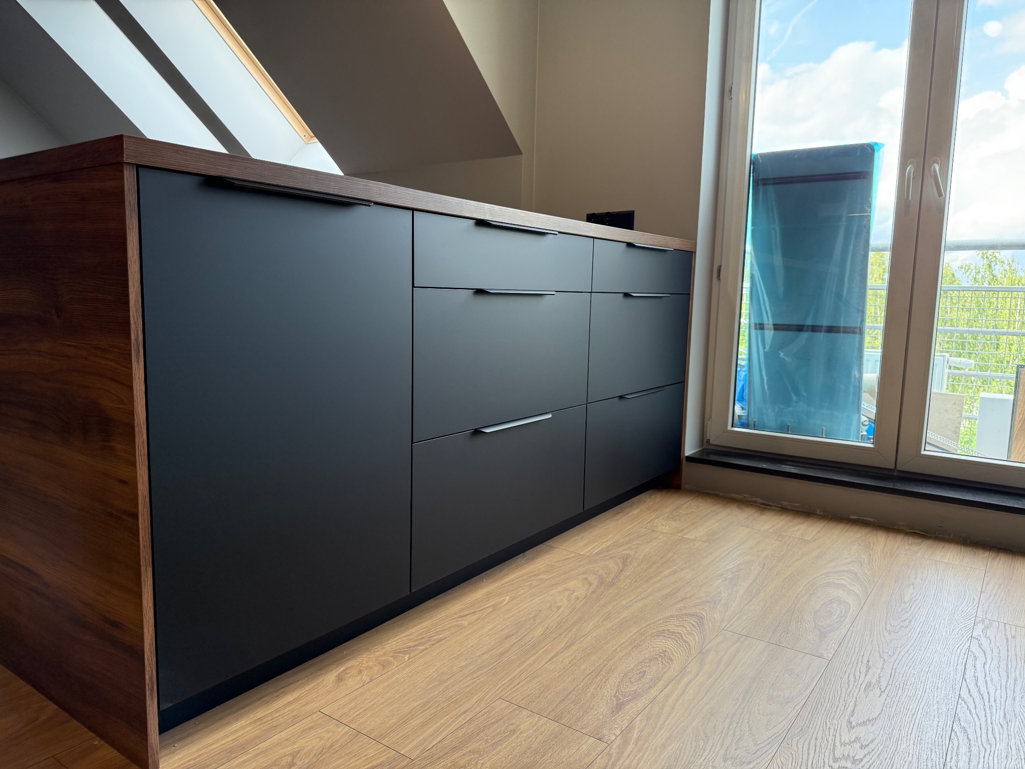 Ciemne, matowe szafki kuchenne z drewnianym blatem w nowoczesnym wnętrzu z oknem balkonowym i podłogą imitującą drewno. Minimalistyczne uchwyty.