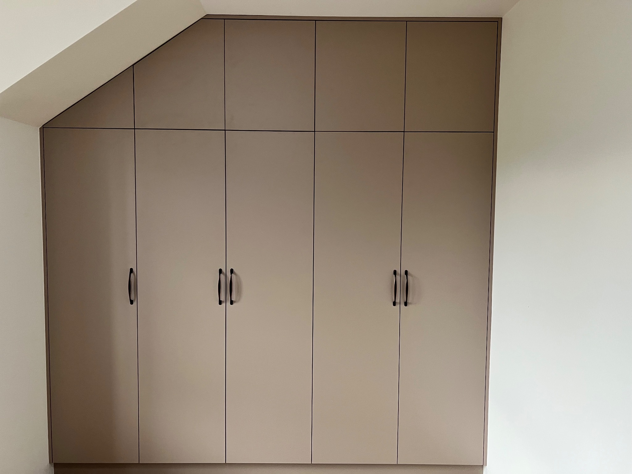 Beżowa szafa wnękowa z czarnymi uchwytami, dopasowana do skosu dachu. Minimalistyczny design, gładkie fronty, zabudowa od podłogi do sufitu.