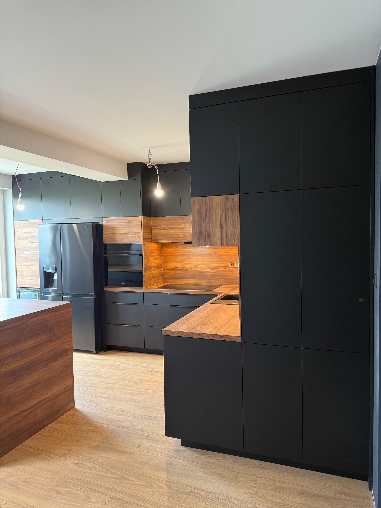 Nowoczesna kuchnia z czarnymi szafkami i drewnianymi akcentami. Widoczny fragment wyspy kuchennej i sprzęt AGD. Oświetlenie punktowe podkreśla detale.