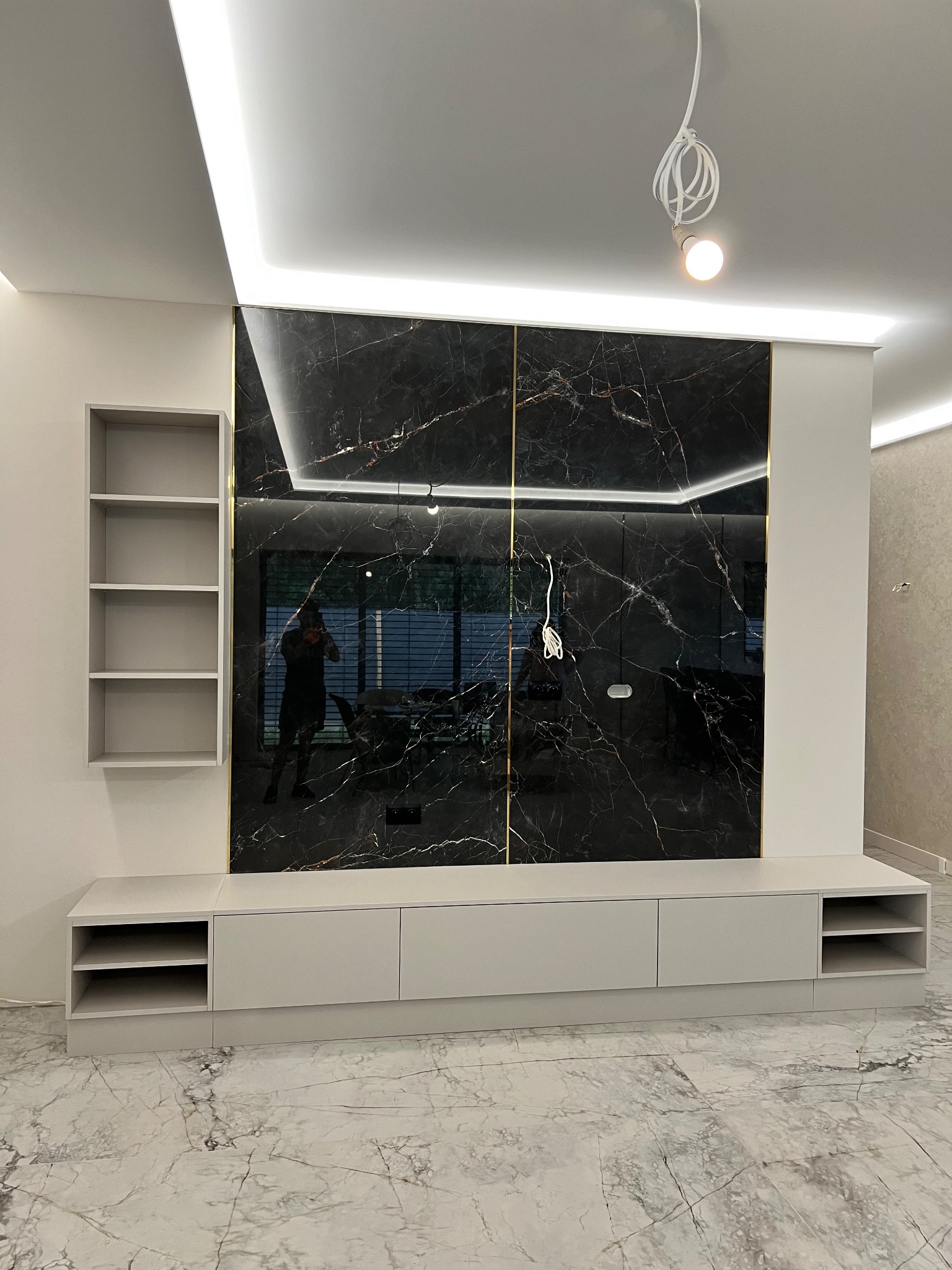 Nowoczesna zabudowa ściany TV z szafkami i półkami w szarości, wykończona czarnym marmurem z złotymi akcentami i podświetleniem LED. Widoczny refleks osoby robiącej zdjęcie.