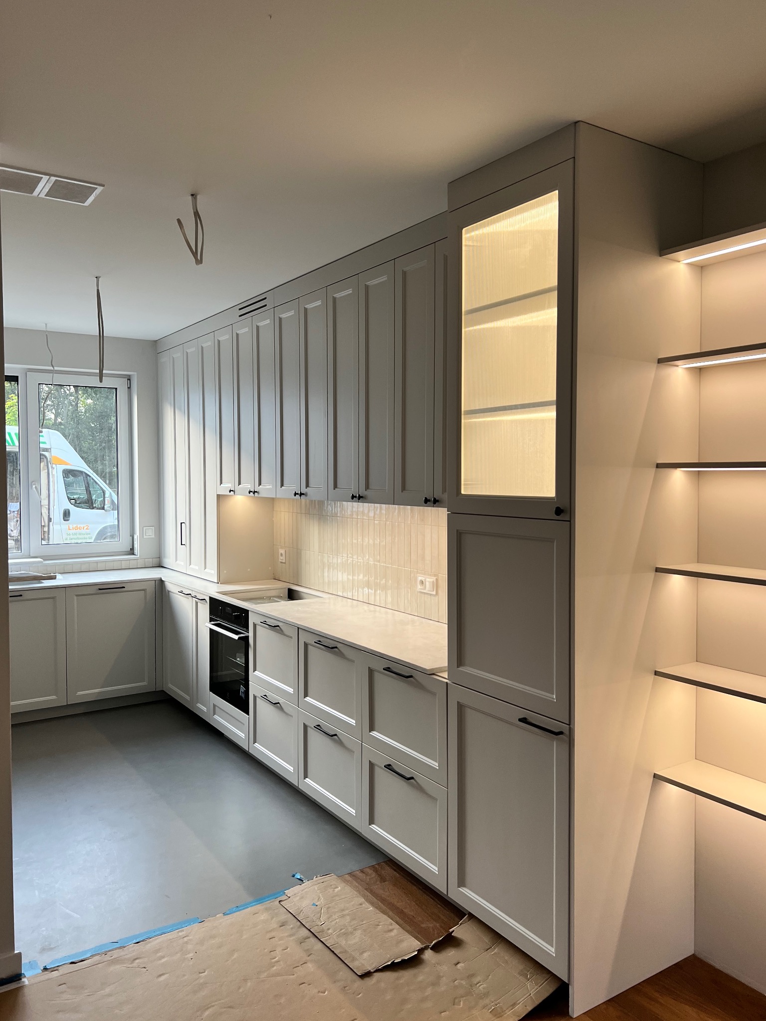 Jasnoszara kuchnia z frezowanymi frontami, czarnymi uchwytami i oświetleniem LED. Widoczne okno i fragment podłogi z zabezpieczeniem na czas montażu.