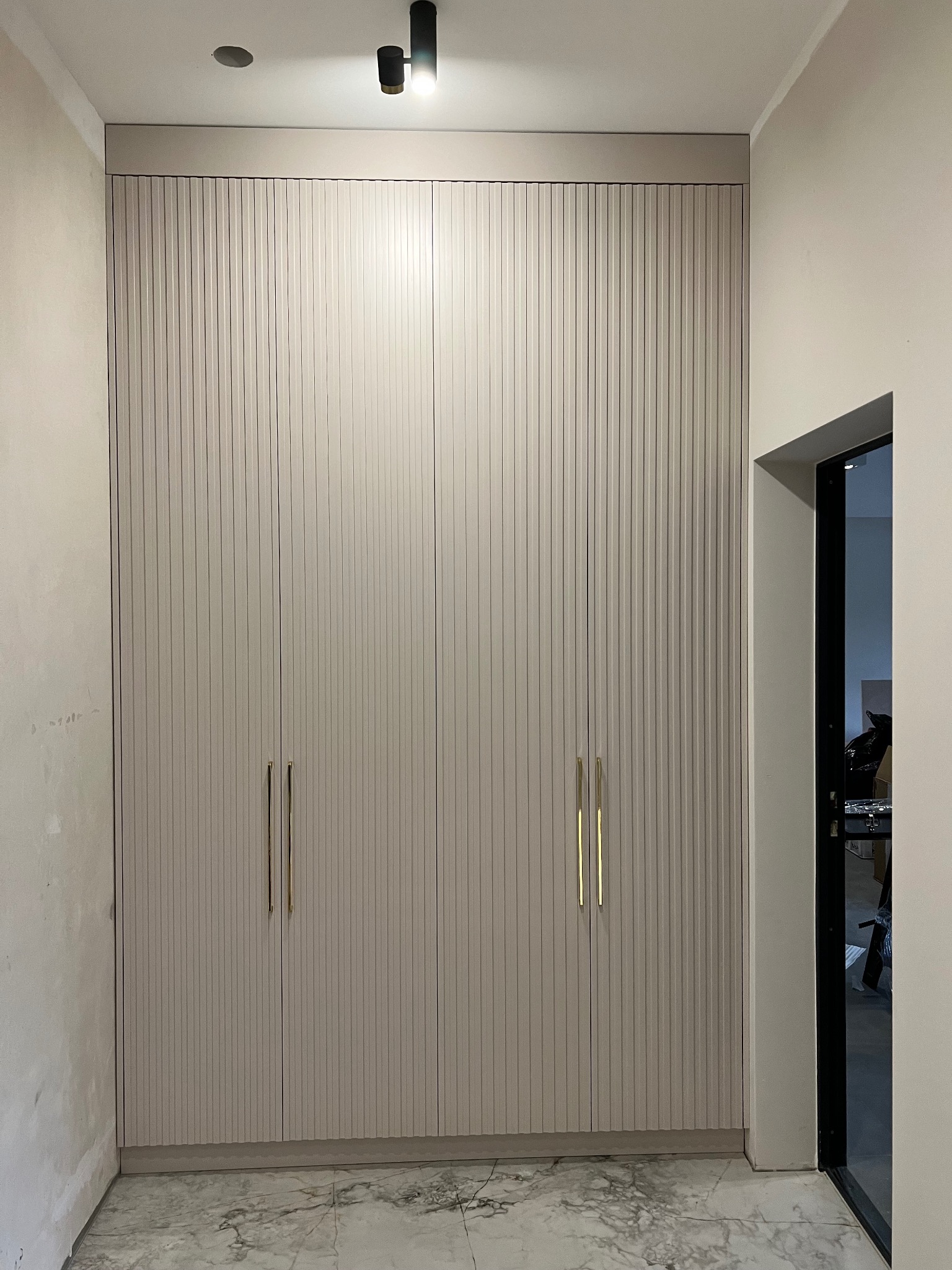 Beżowa szafa wnękowa z ryflowanymi frontami i złotymi uchwytami, dopasowana do wysokości pomieszczenia, widoczna na tle marmurowej podłogi i fragmentu drzwi.