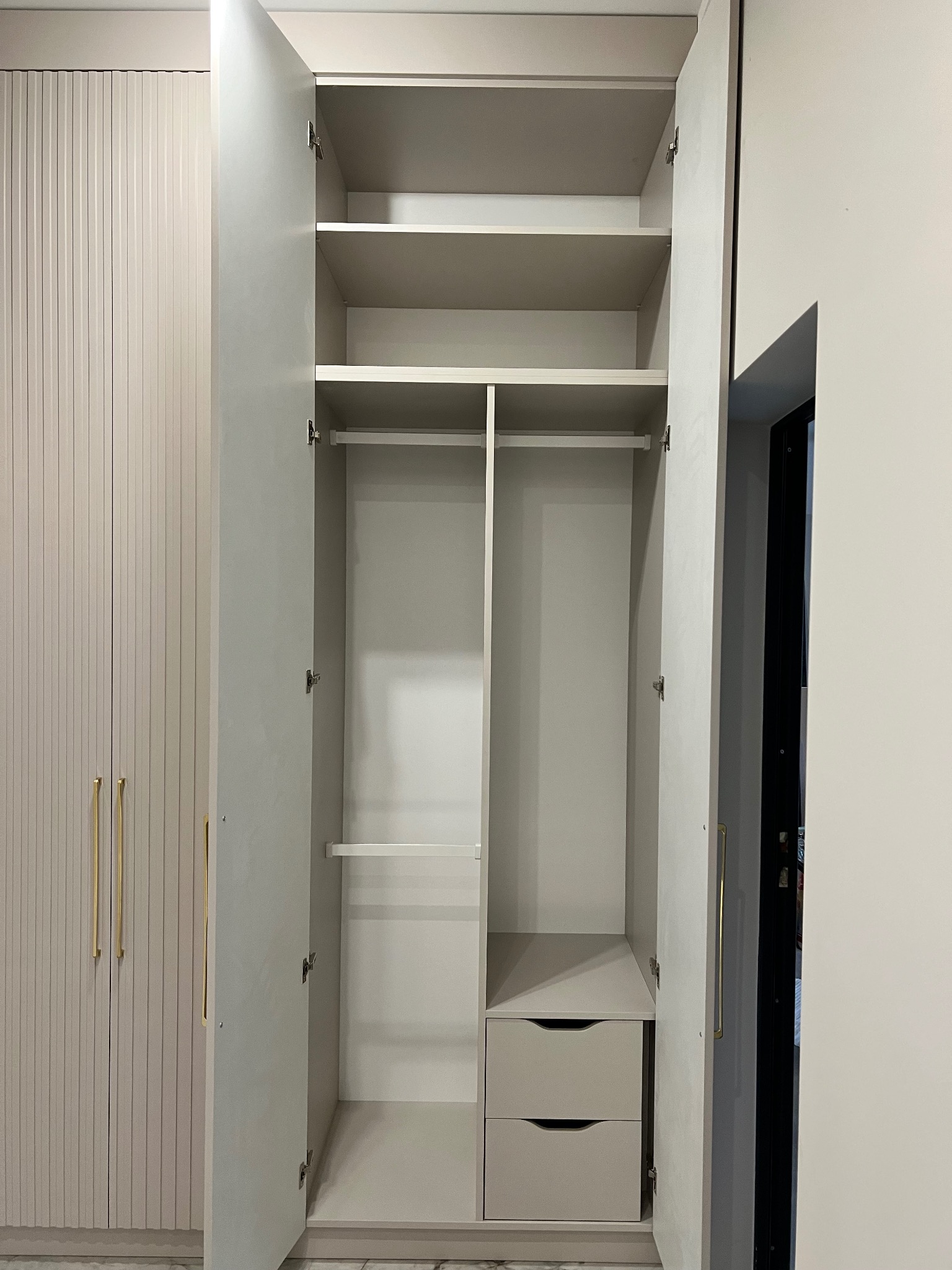 Otwarta szafa wnękowa z szufladami i półkami, fronty w kolorze beżowym z pionowymi frezowaniami, złote uchwyty. Widoczny fragment sąsiedniej szafy.