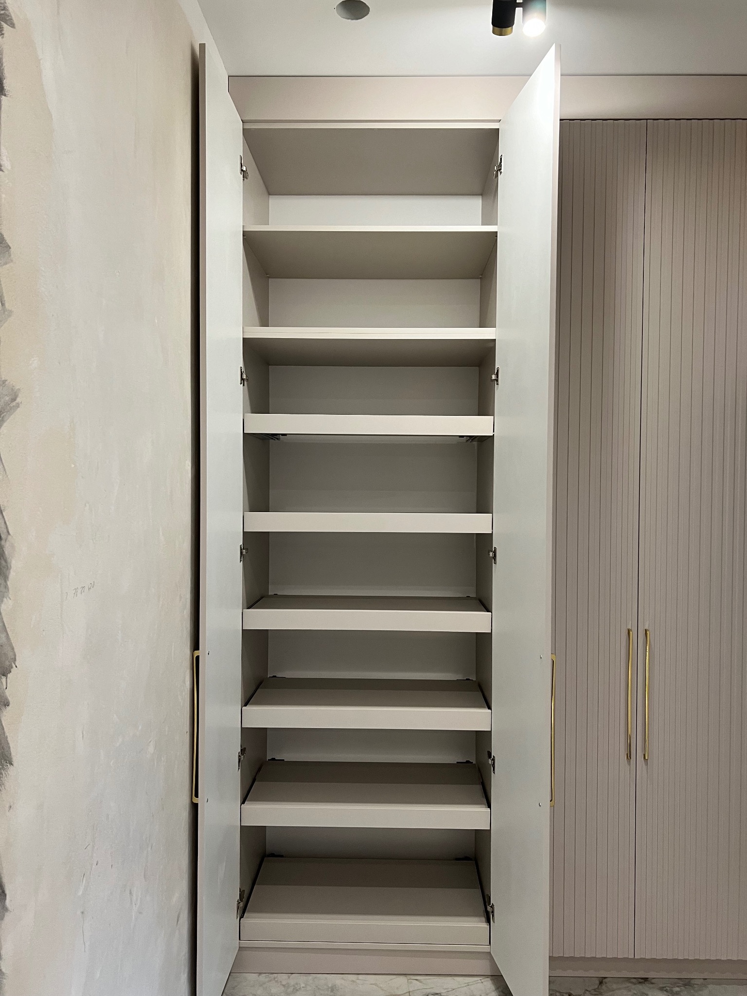 Otwarta, jasna szafa wnękowa z półkami, widok z przodu. Obok szafa z ryflowanymi drzwiami i złotymi uchwytami. Ściana z surowym wykończeniem. Minimalistyczny design.