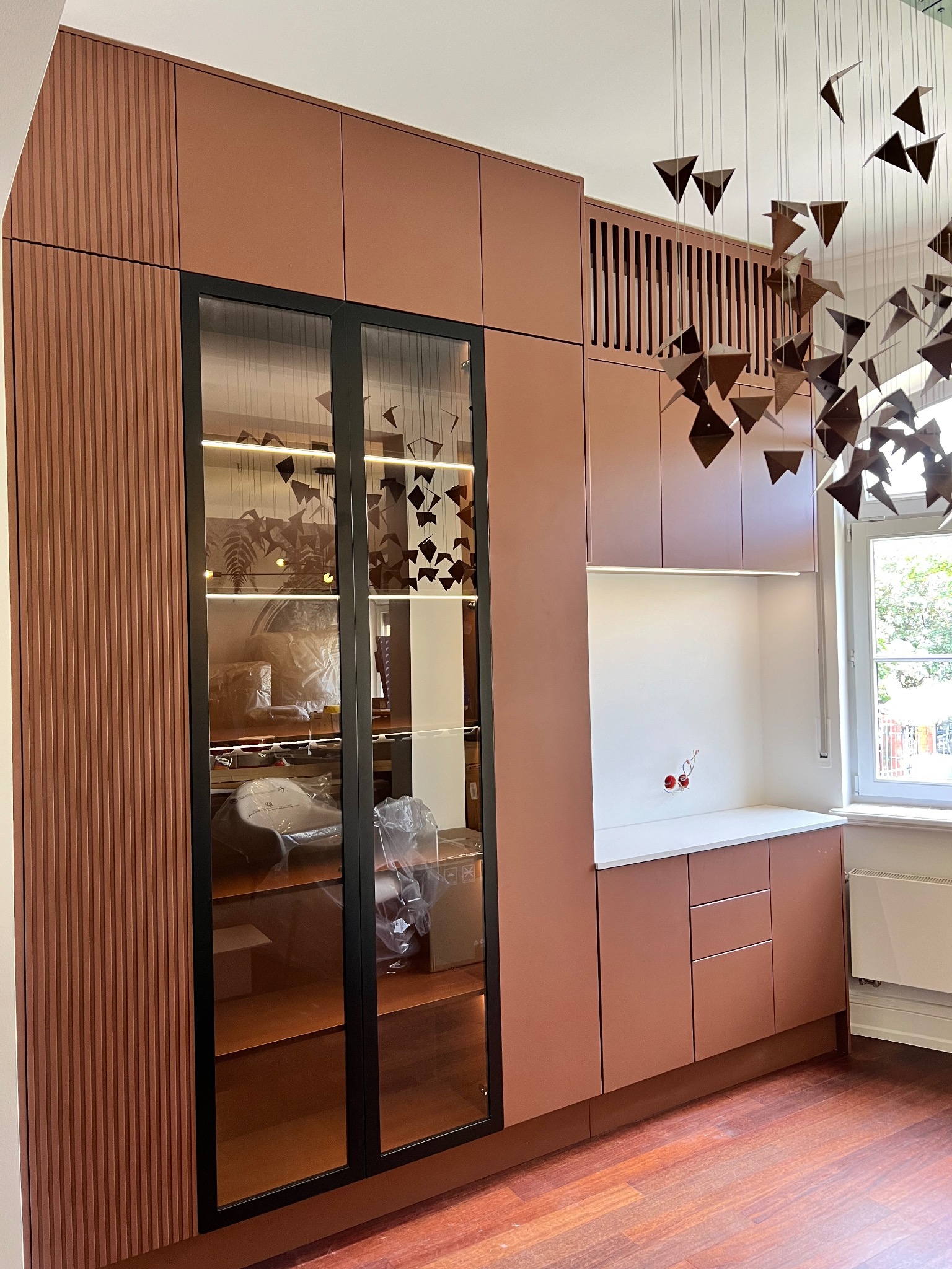 Różowo-brązowa zabudowa meblowa z przeszkloną witryną i dekoracyjnym oświetleniem, uzupełniona wiszącą instalacją artystyczną w nowoczesnym wnętrzu.