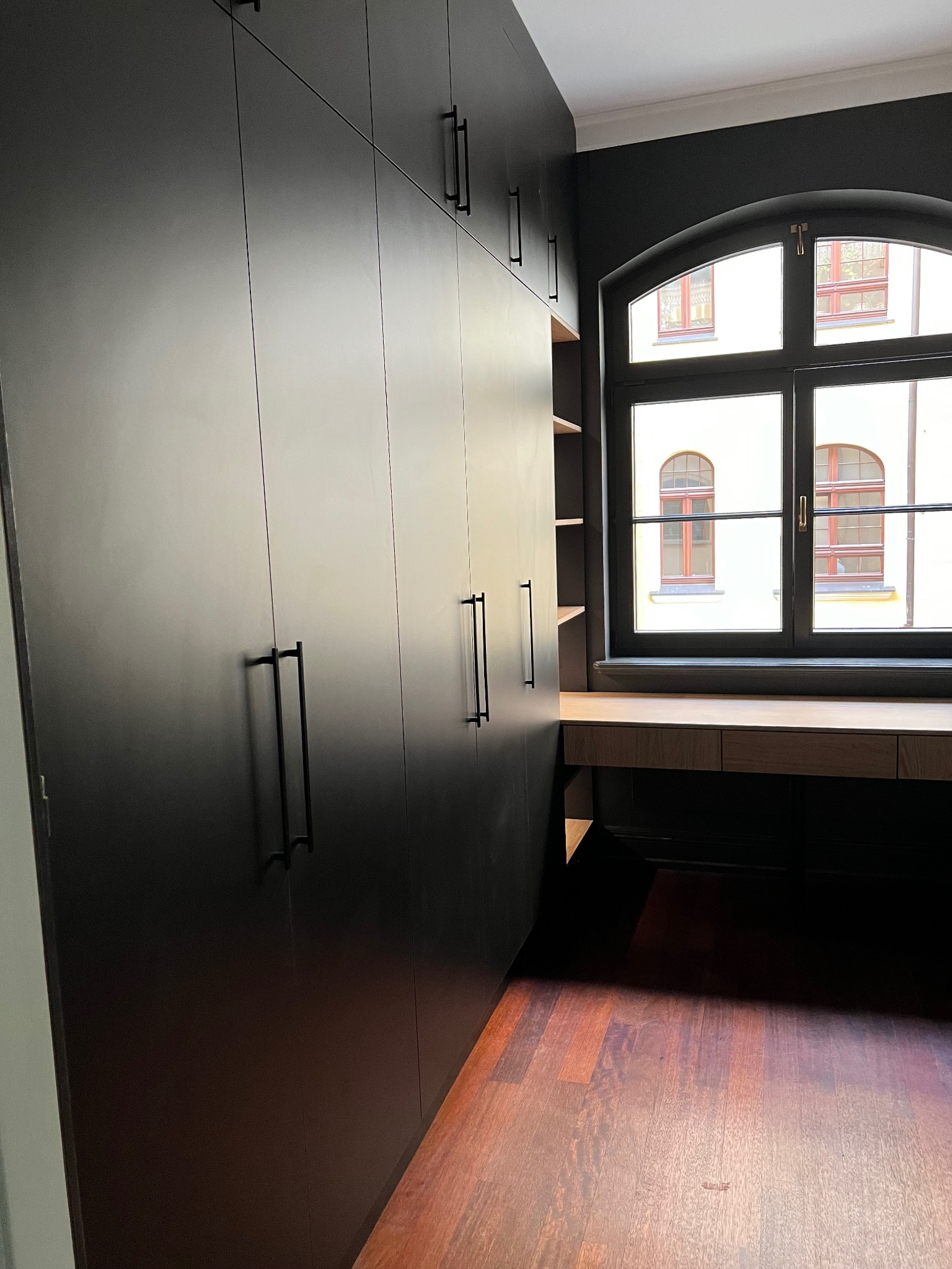 Czarna szafa wnękowa z długimi uchwytami, połączona z biurkiem pod oknem o łukowatym kształcie. Ciemne ściany i drewniana podłoga tworzą elegancki kontrast.