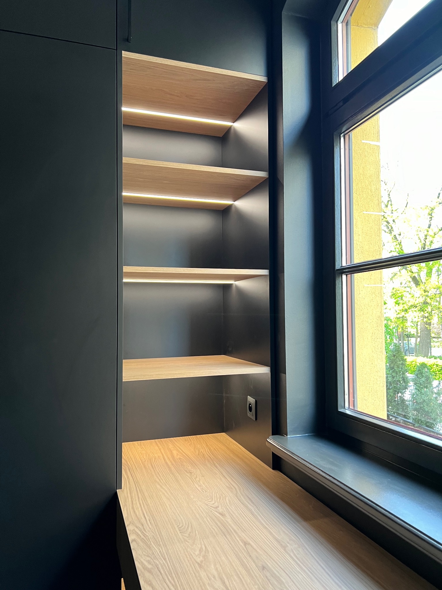 Ciemna szafa wnękowa z drewnianymi półkami podświetlonymi LED, zintegrowana z parapetem okiennym. Nowoczesny design, minimalistyczne wykończenie.