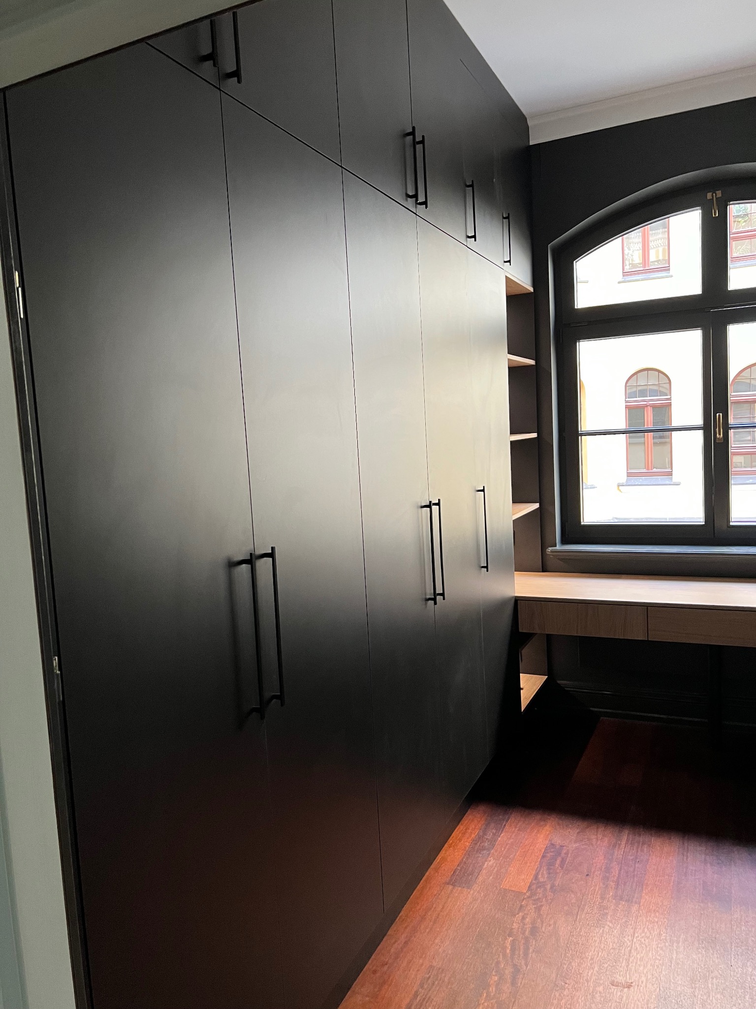 Czarna, matowa szafa wnękowa z długimi uchwytami, dopasowana do biurka pod oknem w kształcie łuku. Parkiet z ciemnego drewna. Minimalistyczny design.