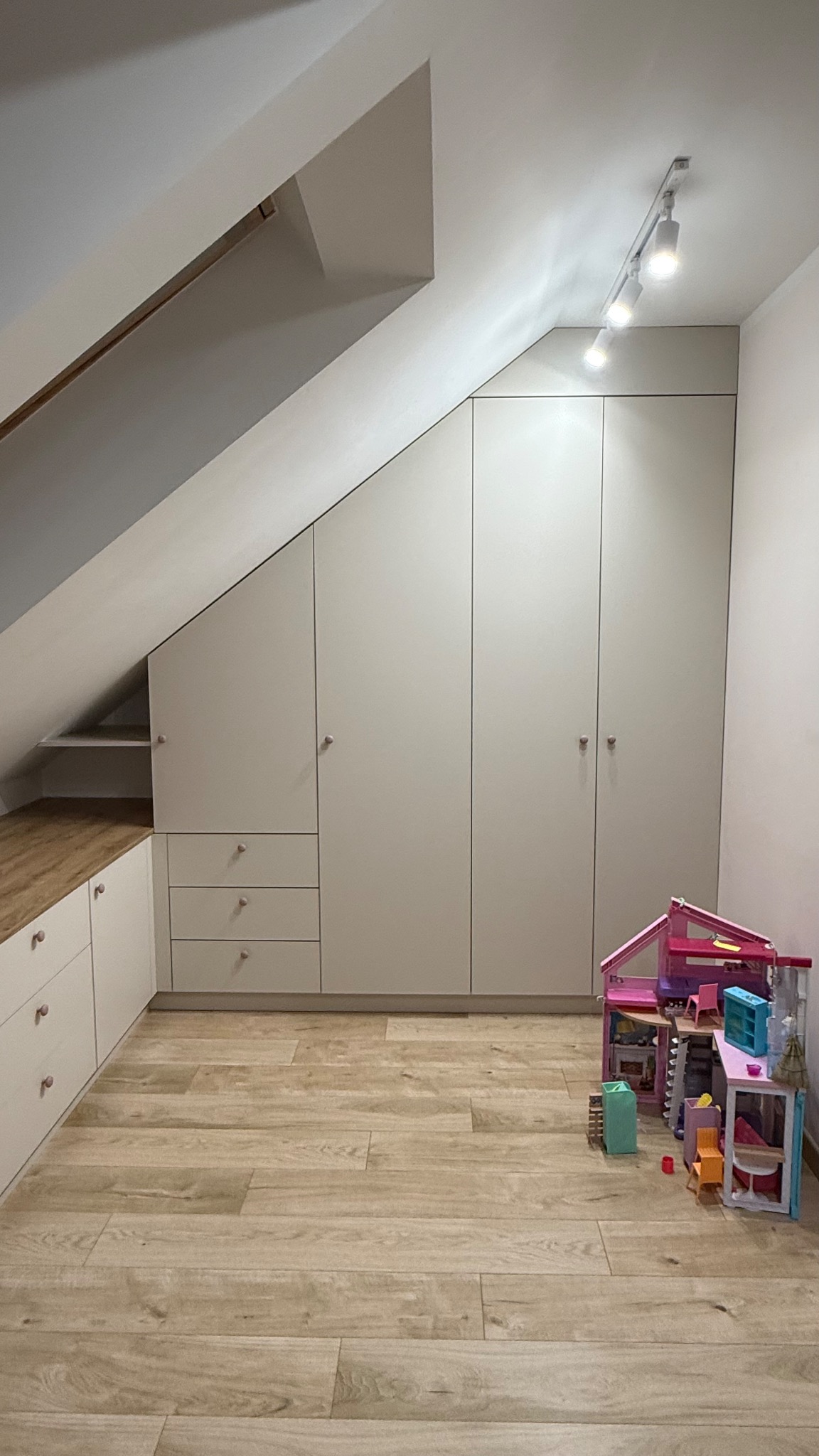 Jasna szafa wnękowa na poddaszu ze skosami, dopasowana do przestrzeni, z drewnianą podłogą i zabawkami dziecięcymi. Oświetlenie punktowe.