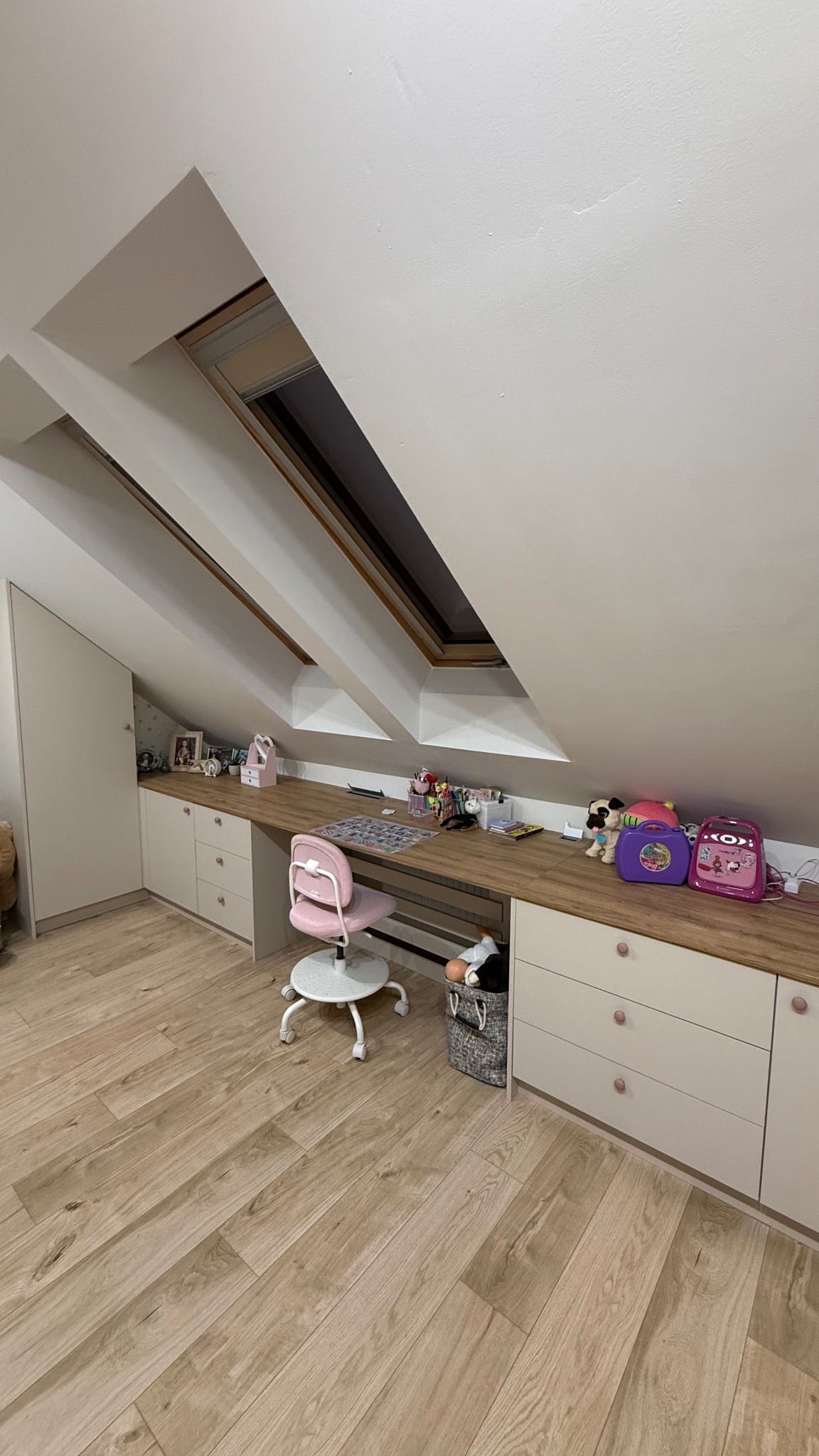 Pokój na poddaszu z zabudową meblową: biurko z blatem drewnopodobnym, białe szafki i szuflady, różowe krzesło obrotowe. Okno dachowe w skosie.