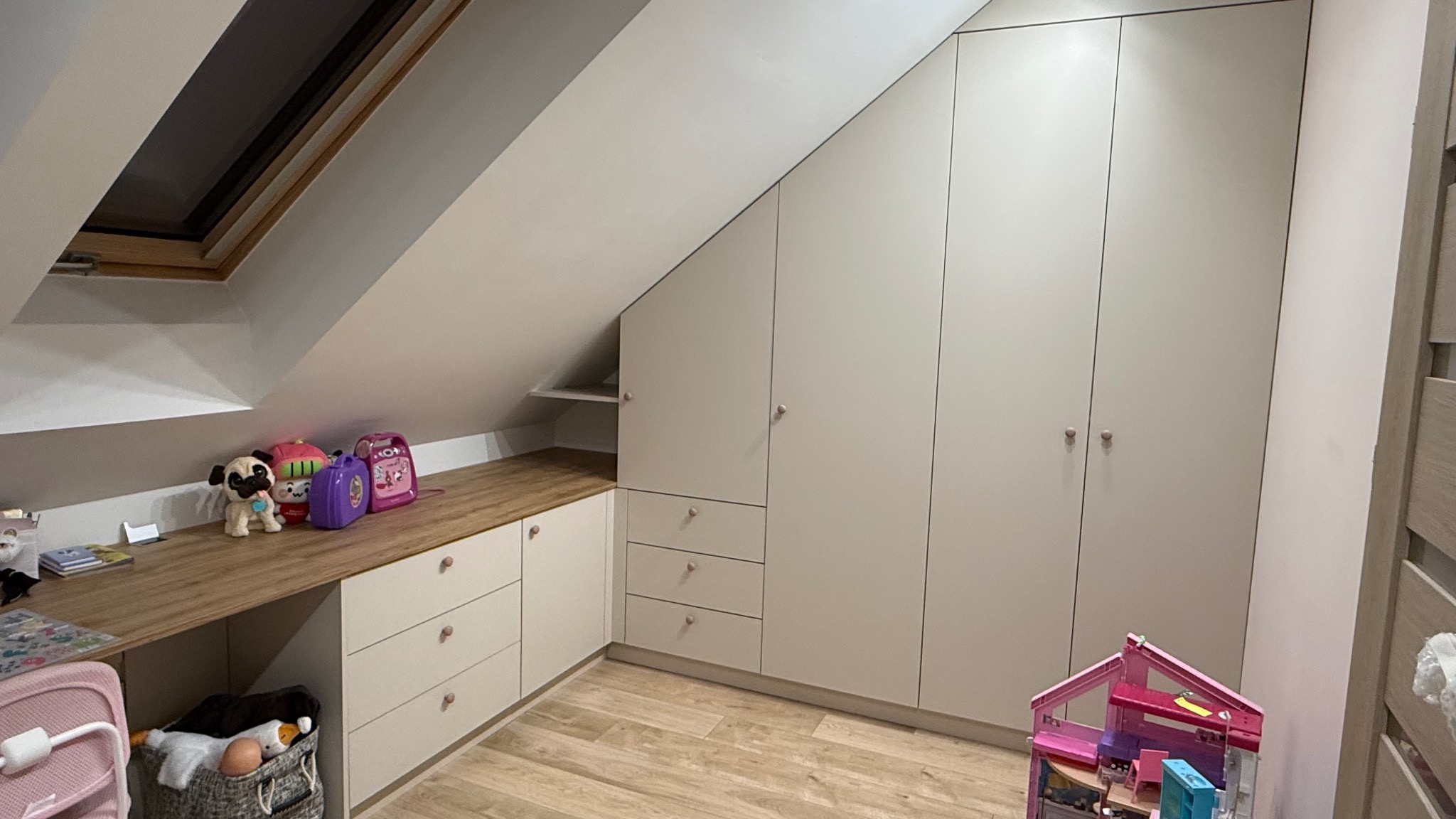 Jasna szafa wnękowa z biurkiem pod skosem na poddaszu, z drewnianym blatem i zabawkami. Ustawna zabudowa meblowa w pokoju dziecięcym.