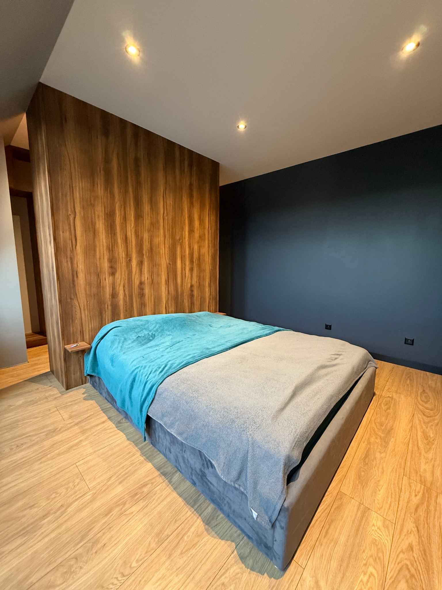 Nowoczesna sypialnia z łóżkiem na podeście, drewnianą ścianą działową i ciemnoniebieską ścianą. Minimalistyczny design, ciepłe oświetlenie punktowe.