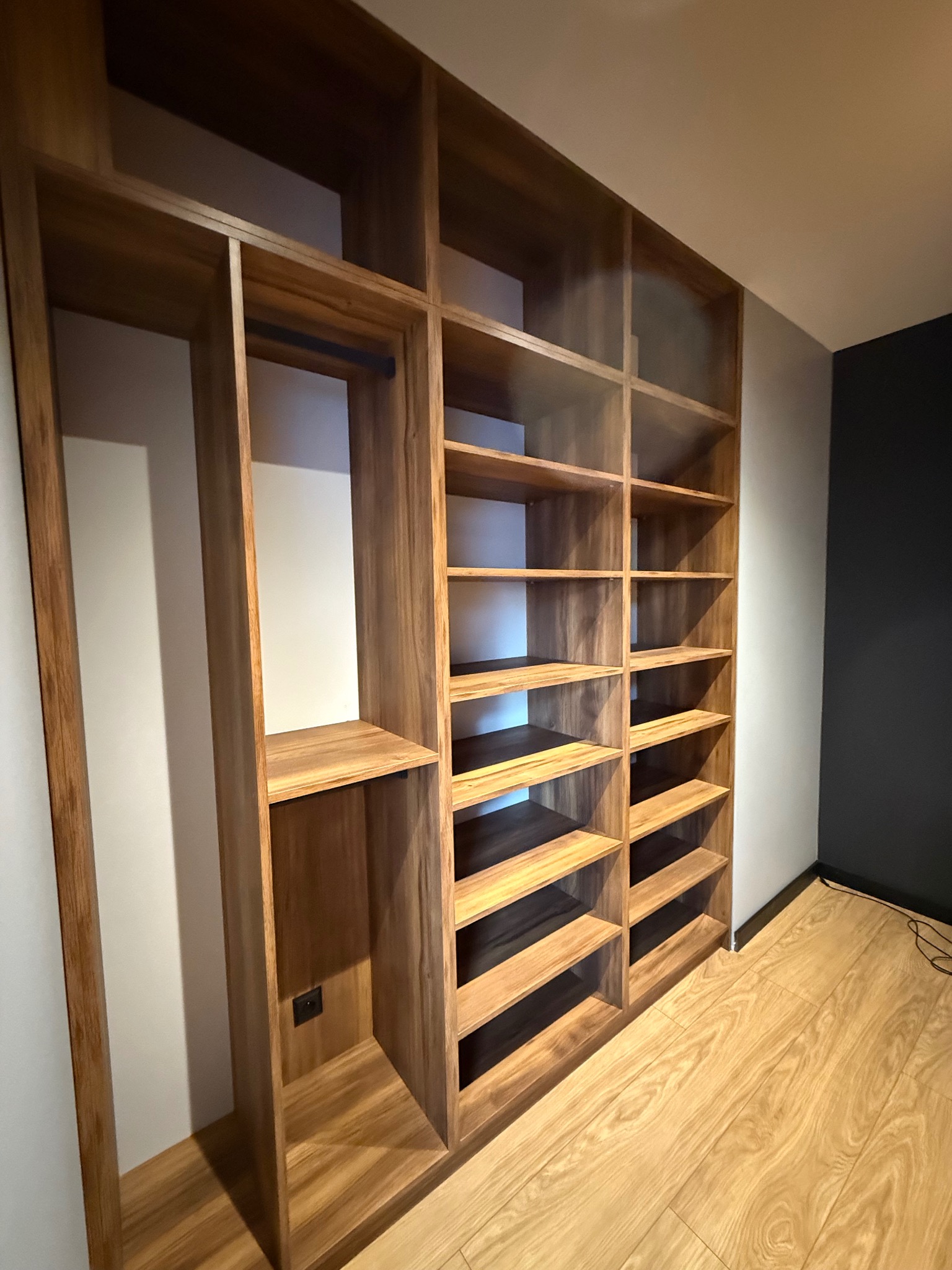 Drewniana szafa wnękowa z półkami i miejscem na wieszaki, z widocznym gniazdkiem elektrycznym, na tle parkietu i ściany w dwóch odcieniach szarości.