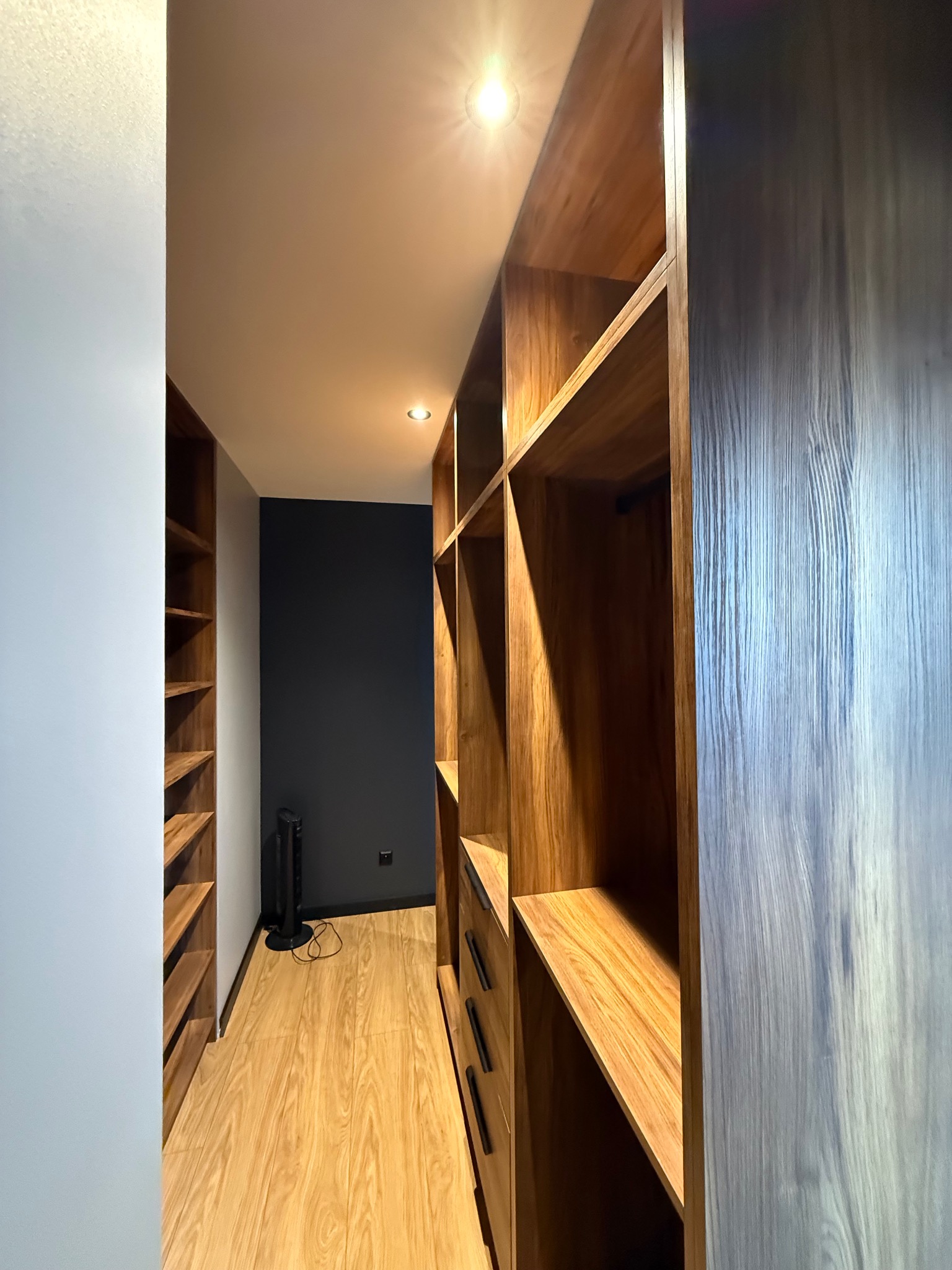 Wnętrze garderoby z drewnianymi półkami i szufladami, oświetlone punktowo, z ciemną ścianą w tle i drewnianą podłogą. Widok z perspektywy wejścia.