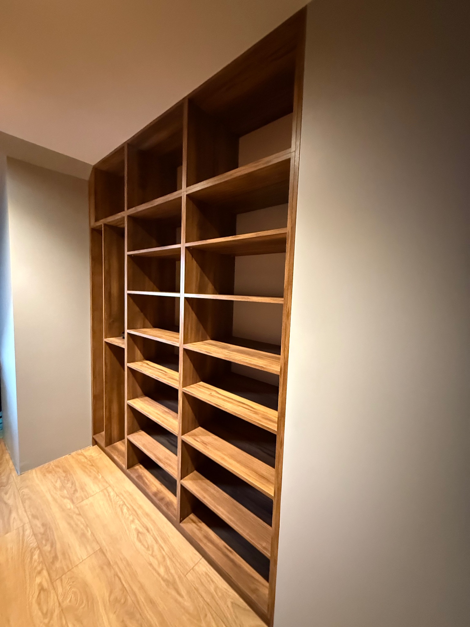 Drewniana szafa wnękowa z półkami, dopasowana do przestrzeni, z widoczną strukturą drewna i oświetleniem, wkomponowana w jasne wnętrze.