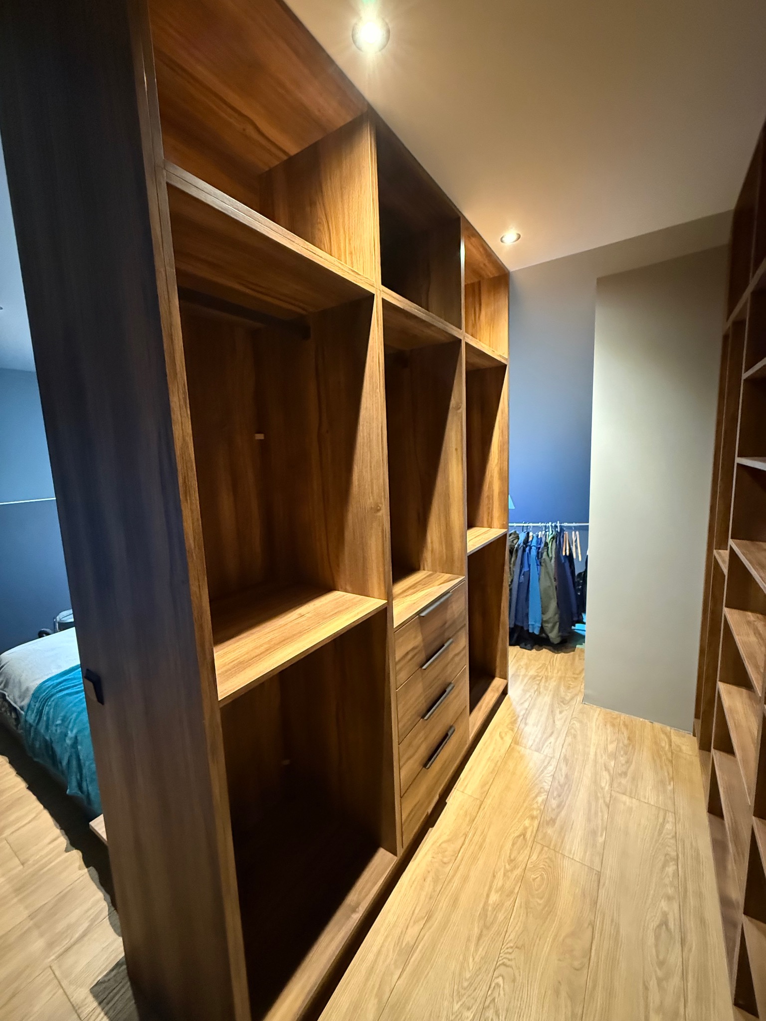 Drewniana szafa wnękowa z półkami i szufladami, widok z perspektywy wnętrza, oświetlenie punktowe, podłoga z paneli, w tle widoczne ubrania na wieszakach.