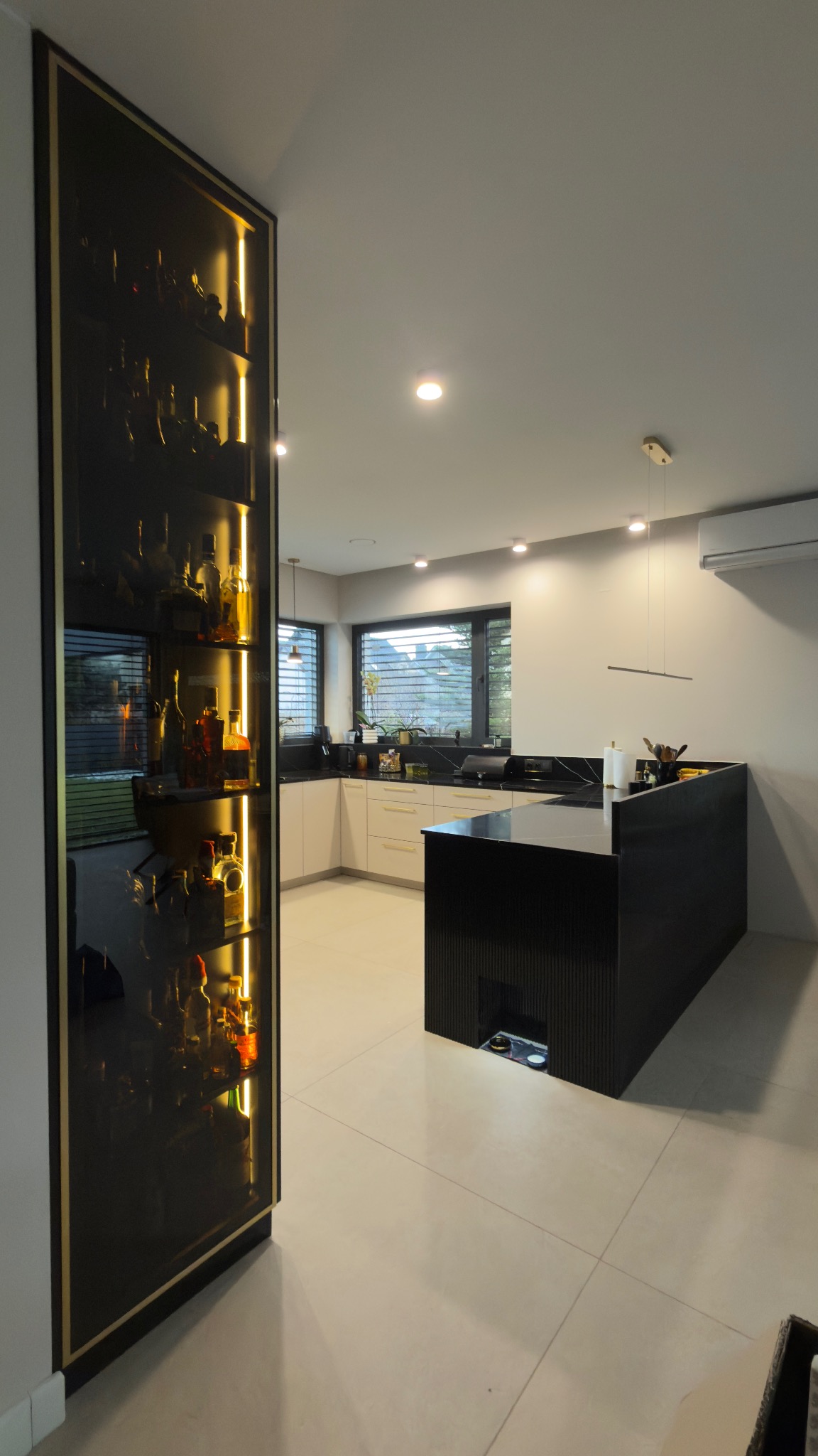 Nowoczesna kuchnia z wyspą i podświetlaną szafką na alkohole. Białe fronty szafek, czarny blat i wyspa. Minimalistyczne oświetlenie.