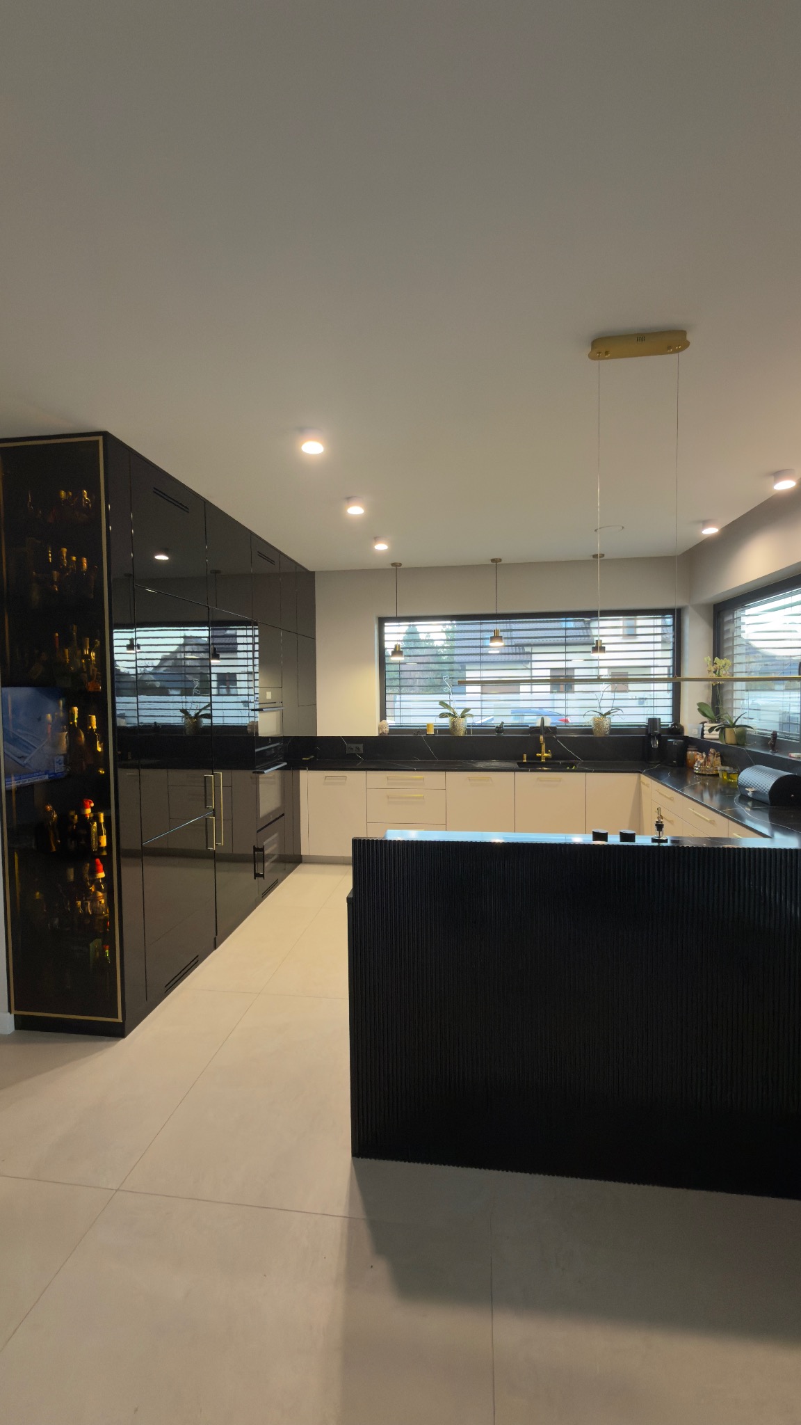 Nowoczesna kuchnia z czarnymi, lakierowanymi frontami i złotymi akcentami, wyspa z ryflowanym wykończeniem, oświetlenie punktowe i okno z żaluzjami.