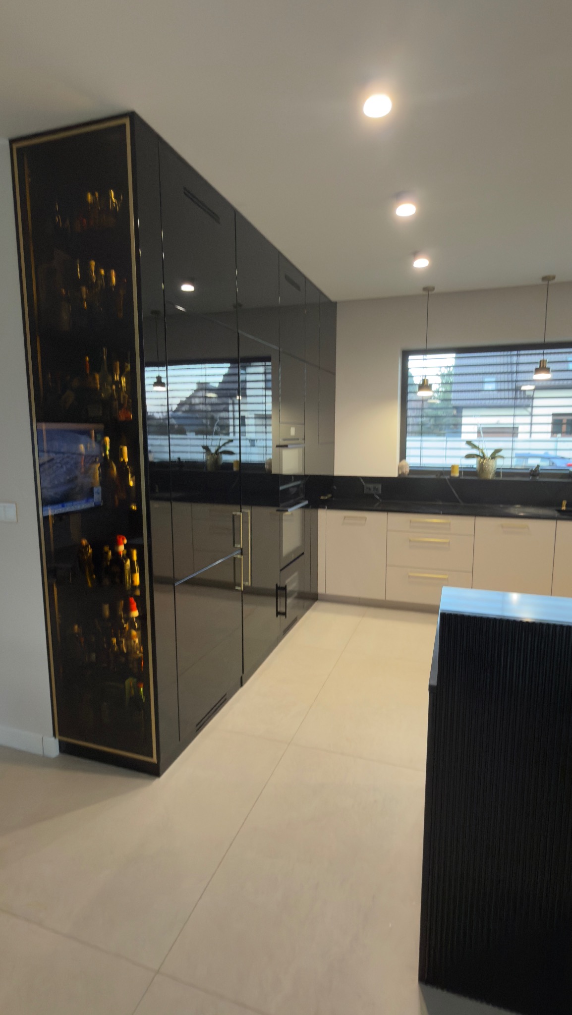 Nowoczesna kuchnia z czarnymi, lakierowanymi frontami i złotymi akcentami, przeszklona witryna z oświetleniem, jasne blaty i płytki podłogowe, okno z żaluzjami.