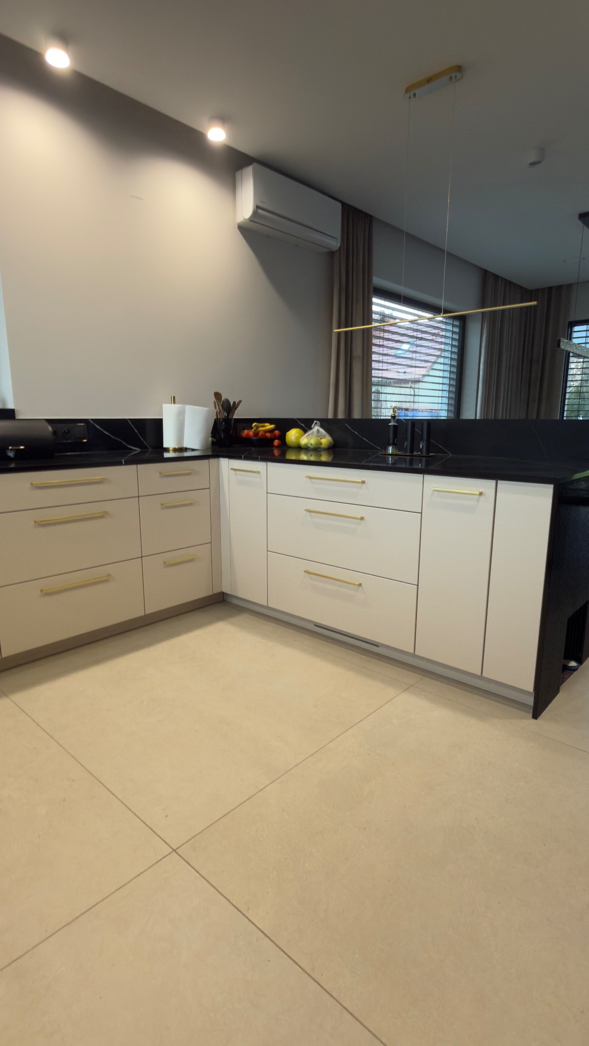 Jasna kuchnia z czarnym blatem i złotymi uchwytami, oświetlona nowoczesną lampą. Na blacie widoczne owoce i akcesoria kuchenne. Minimalistyczny design.