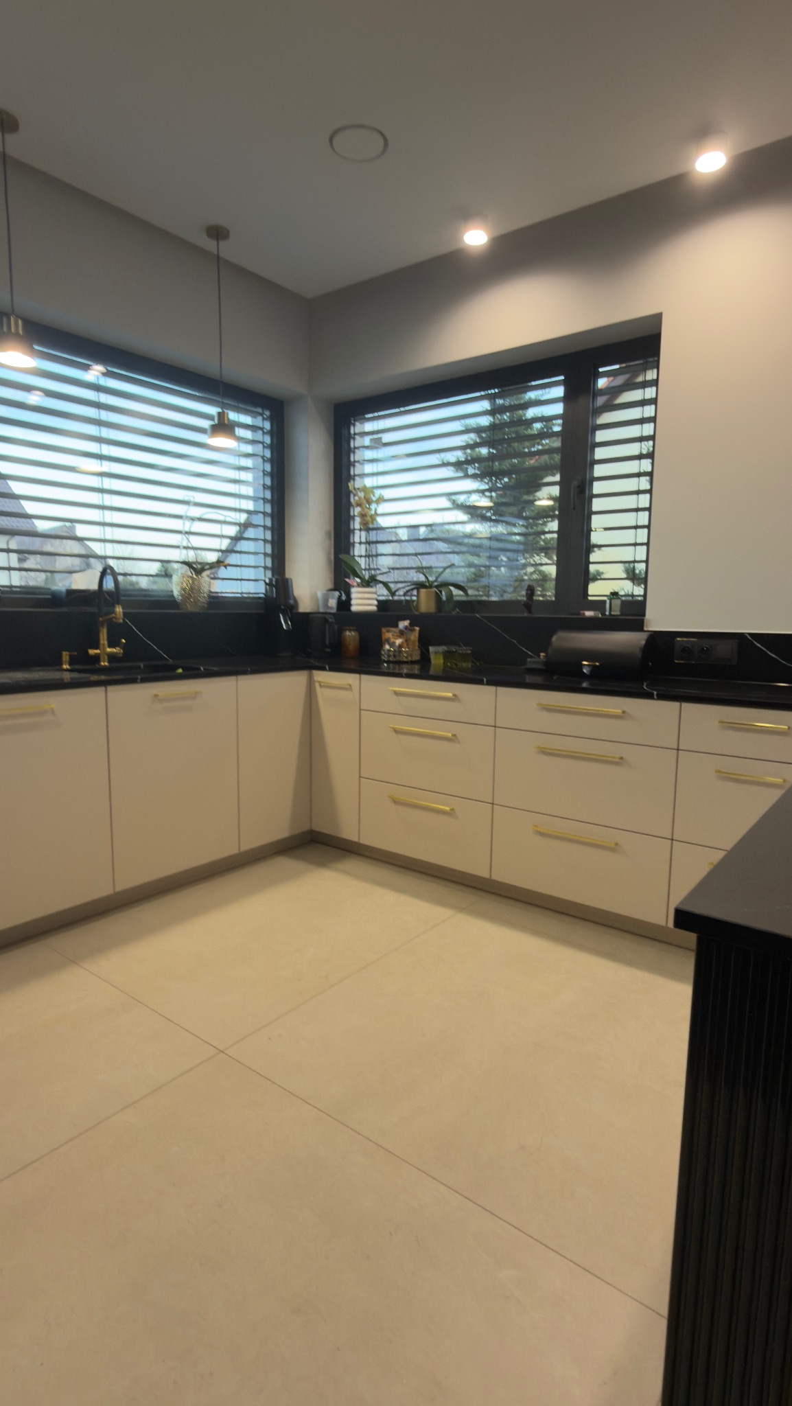 Jasna kuchnia z czarnym blatem i złotymi uchwytami, widok na okno z żaluzjami. Minimalistyczny design z elementami dekoracyjnymi.