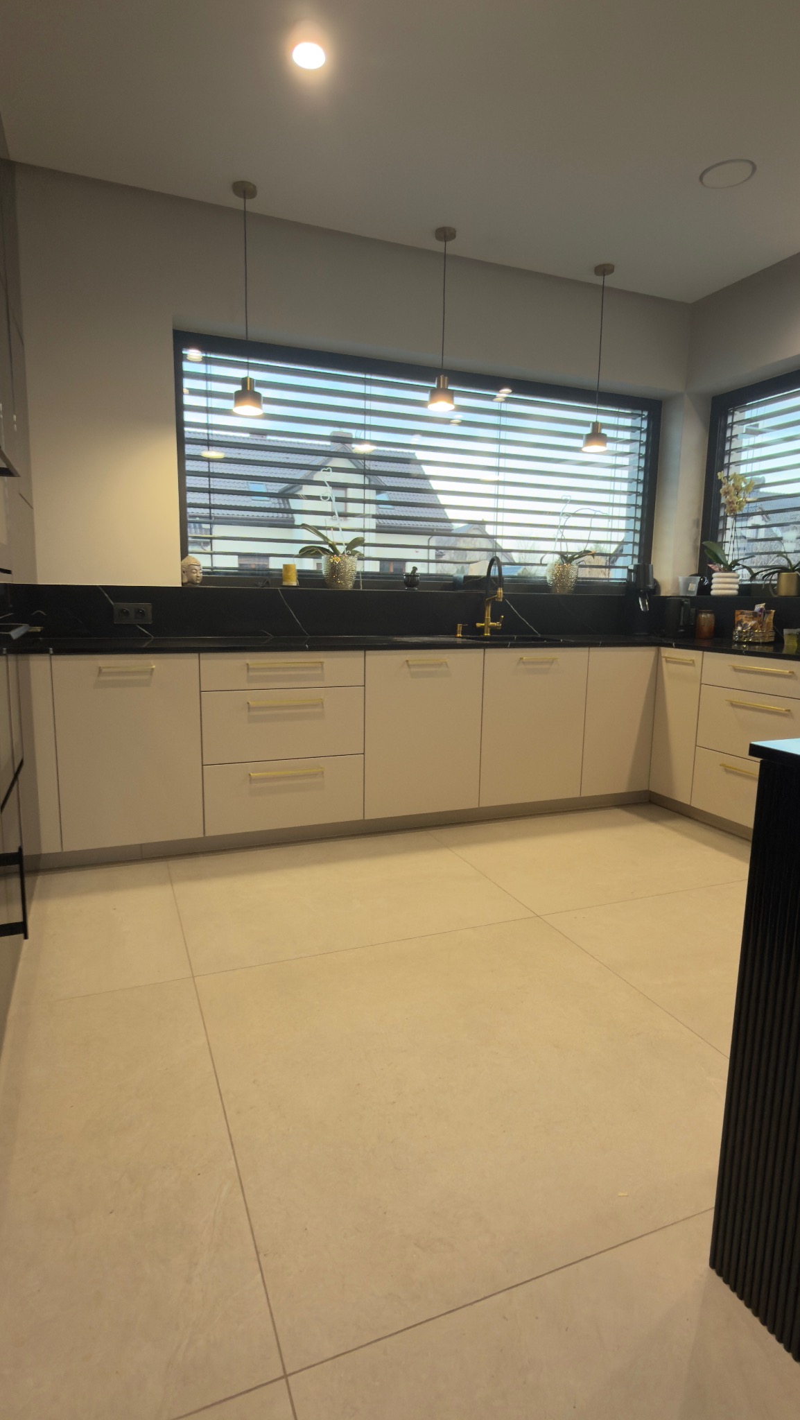 Jasna kuchnia z czarnym blatem i złotymi akcentami, oświetlona wiszącymi lampami. Okno z żaluzjami w tle. Minimalistyczny design.