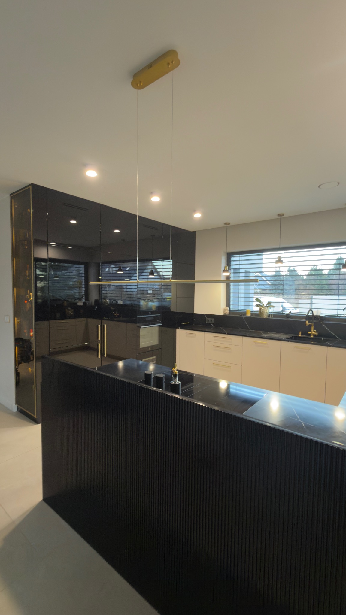 Nowoczesna kuchnia z wyspą: czarne, ryflowane fronty wyspy, jasne szafki, czarny blat z marmurowym wzorem, złote akcenty oświetlenia i armatury, lustrzane szafki.