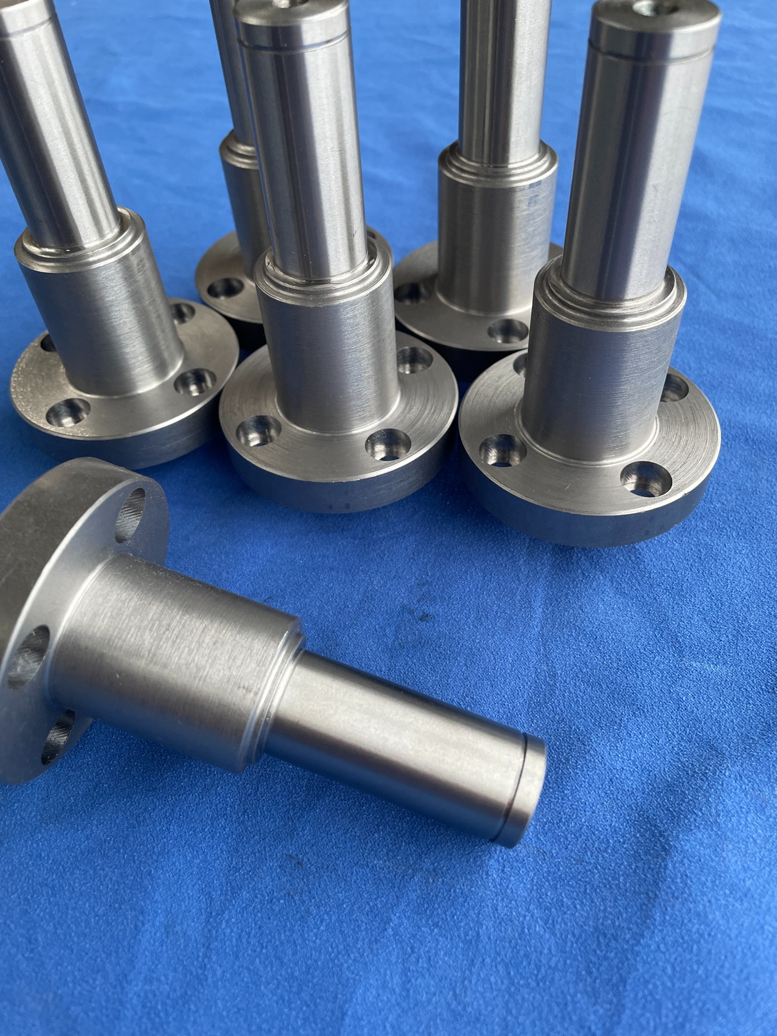 Srebrne, metalowe elementy z kołnierzami i otworami na niebieskim tle. Precyzyjnie wykonane detale, widoczne ślady obróbki. Elementy gotowe do montażu.