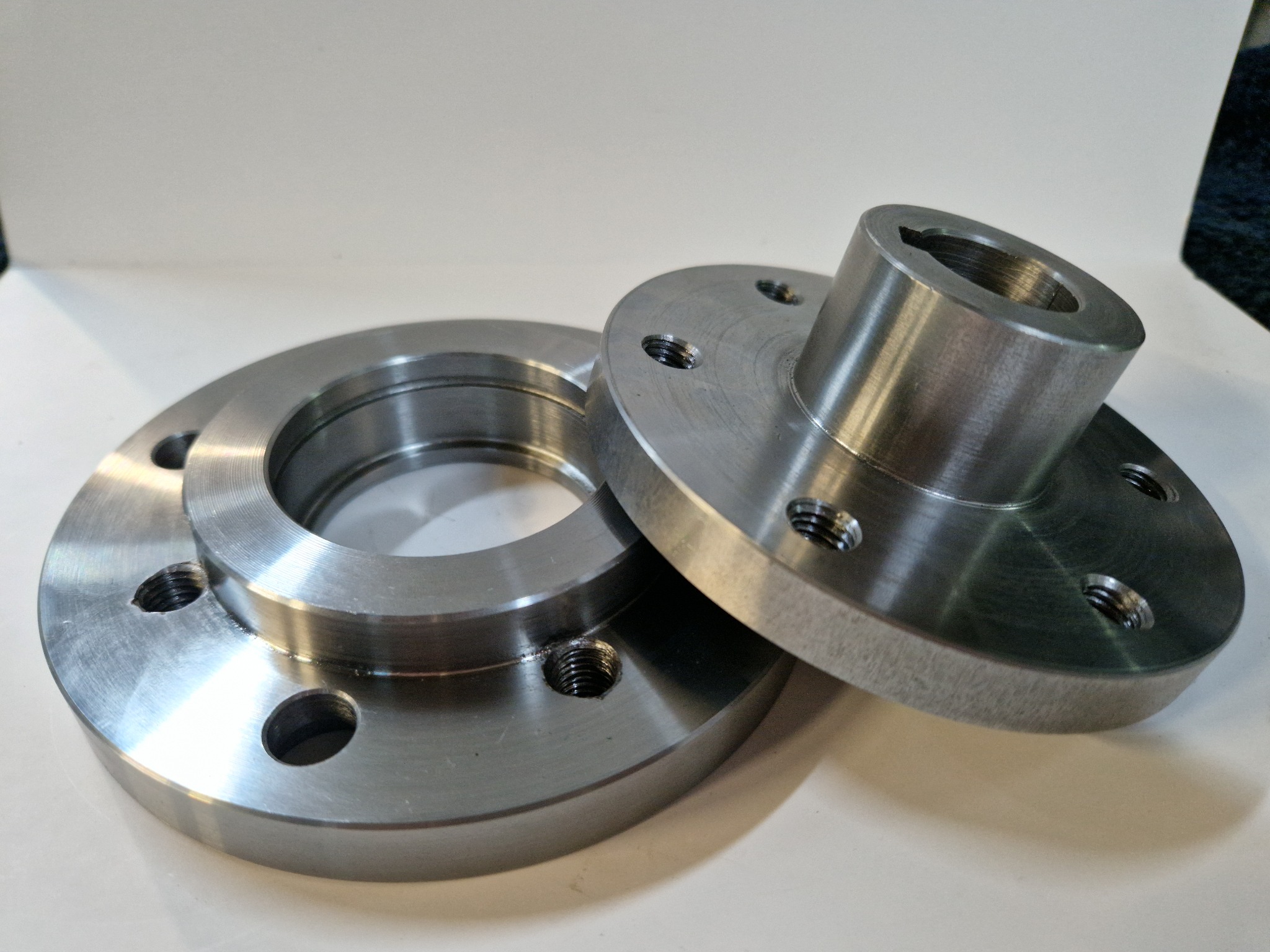 Dwie precyzyjnie wykonane, metalowe flansze z otworami na śruby, leżące na białym tle. Widoczne ślady obróbki skrawaniem i gwintowane otwory.