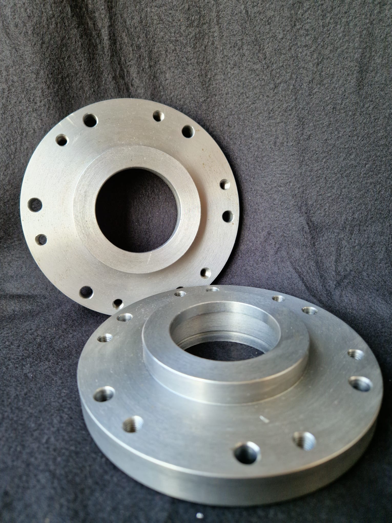 Dwie srebrne, metalowe flansze z otworami na śruby, ułożone na ciemnoszarym tle. Widoczne ślady obróbki skrawaniem i delikatne refleksy świetlne.