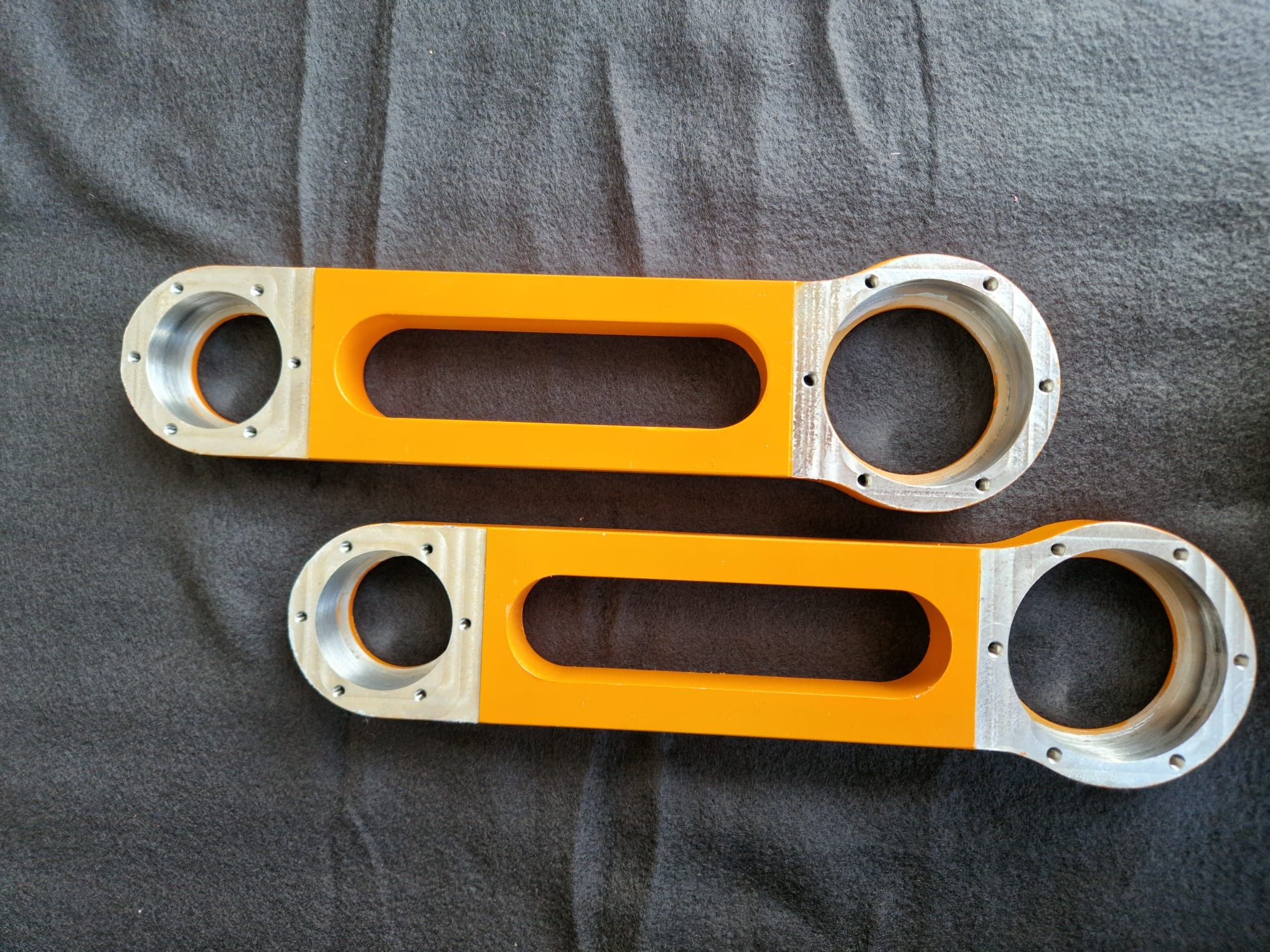 Dwa pomarańczowe, metalowe elementy z otworami na szarym tle. Elementy są identyczne, ułożone równolegle, z widocznymi śladami obróbki.