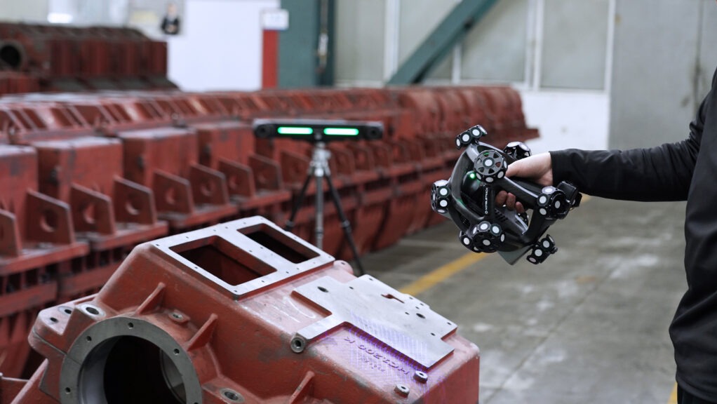 Skanowanie 3D czerwonej, metalowej obudowy maszyny za pomocą ręcznego skanera. W tle widoczny skaner stacjonarny na statywie i rząd podobnych obudów.