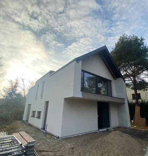 Nowoczesny, dwukondygnacyjny dom z białym tynkiem, dużym oknem i ciemnym dachem. Widoczne rusztowania i piasek wskazują na trwające prace wykończeniowe.