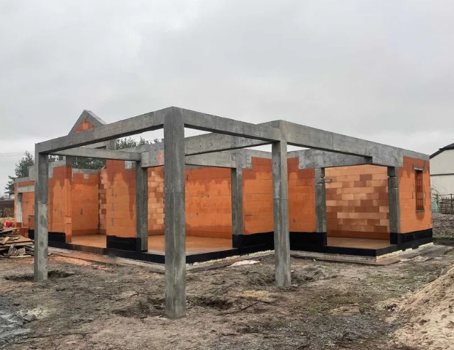 Surowy stan domu z czerwonej cegły z żelbetową konstrukcją szkieletową, widok z zewnątrz na tle pochmurnego nieba. Fundament zabezpieczony izolacją.