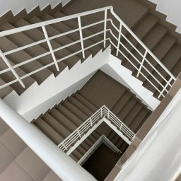 NOWE GORYZONTY SPÓŁKA Z OGRANICZONĄ ODPOWIEDZIALNOŚCIĄ - Klatka schodowa z brązowymi schodami i białymi barierkami, widziana z góry w geometrycznym ujęciu. Minimalistyczny design, symetryczne linie i neutralna kolorystyka.