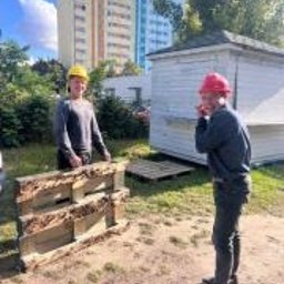 NOWE GORYZONTY SPÓŁKA Z OGRANICZONĄ ODPOWIEDZIALNOŚCIĄ - Dwóch mężczyzn w kaskach, jeden żółty, drugi czerwony, stoi obok drewnianych skrzyń na tle budynku i zieleni. Słoneczny dzień, widoczne cienie.