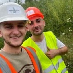 NOWE GORYZONTY SPÓŁKA Z OGRANICZONĄ ODPOWIEDZIALNOŚCIĄ - Uśmiechnięci pracownicy w kaskach i kamizelkach odblaskowych na tle zieleni. Selfie z budowy, widoczne logo firmy na kasku.
