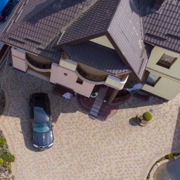 NOWE GORYZONTY SPÓŁKA Z OGRANICZONĄ ODPOWIEDZIALNOŚCIĄ - Widok z góry na posesję z beżowo-brązową kostką brukową, ciemny samochód na podjeździe, dom z balkonami i brązowym dachem, mały staw i zadbana zieleń.