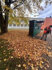 Jesienne sprzątanie: mężczyzna w kamizelce odblaskowej grabi liście obok zielonego kontenera na tle budynku z oknami i drzewa z żółtymi liśćmi.