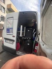 Wnętrze białego busa z otwartymi drzwiami, wypełnione sprzętem i torbami, prawdopodobnie związanym z usługami sprzątania lub remontowymi. Widoczny fragment budynku w tle.