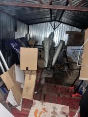 Wnętrze blaszaka z chaotycznie ułożonymi kartonami, rurami i innymi przedmiotami. Widoczne metalowe ściany i sufit. Podłoga z czerwonej cegły.