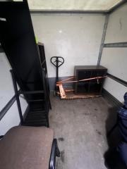 Wnętrze furgonetki z meblami zabezpieczonymi pomarańczową taśmą na palecie, w tle wózek paletowy. Widoczne elementy wyposażenia wnętrza pojazdu.