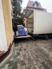 Rozładunek palety z farbami z samochodu dostawczego przy budynku z ceglaną elewacją. Widoczny fragment elewacji i kostka brukowa.