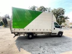 Biały samochód dostawczy z zielono-białą plandeką na parkingu w słoneczny dzień. Widoczny cień pojazdu i budynki w tle. Charakterystyczny wzór na plandece.