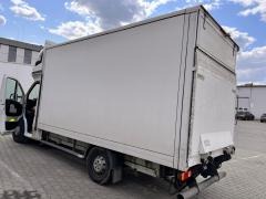 Biały samochód dostawczy z otwartymi drzwiami, widok z tyłu i boku, na tle budynku przemysłowego. Widoczne elementy podwozia i opony.