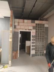 Wnętrze w trakcie prac murarskich: częściowo wymurowana ściana z cegieł i luksferów, widoczny fragment sufitu i posadzki, osoba w tle. Etap budowy ścianki działowej.