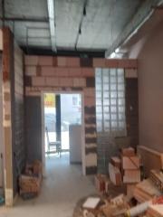 Wnętrze w trakcie prac murarskich: częściowo wymurowana ściana z otworem drzwiowym, bloczki i narzędzia budowlane w pomieszczeniu z surowym wykończeniem.