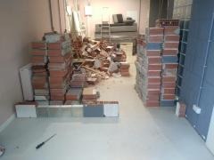 Rozbiórka ściany z cegły w pomieszczeniu. Widoczne gruz, cegły ułożone w stosy, biała podłoga i fragment instalacji elektrycznej. Etap prac rozbiórkowych.