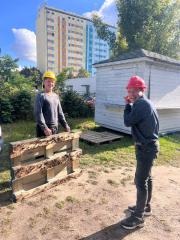 Dwóch mężczyzn w kaskach, jeden żółty, drugi czerwony, stoi obok drewnianych skrzyń na tle budynku i zieleni. Słoneczny dzień, widoczne cienie.